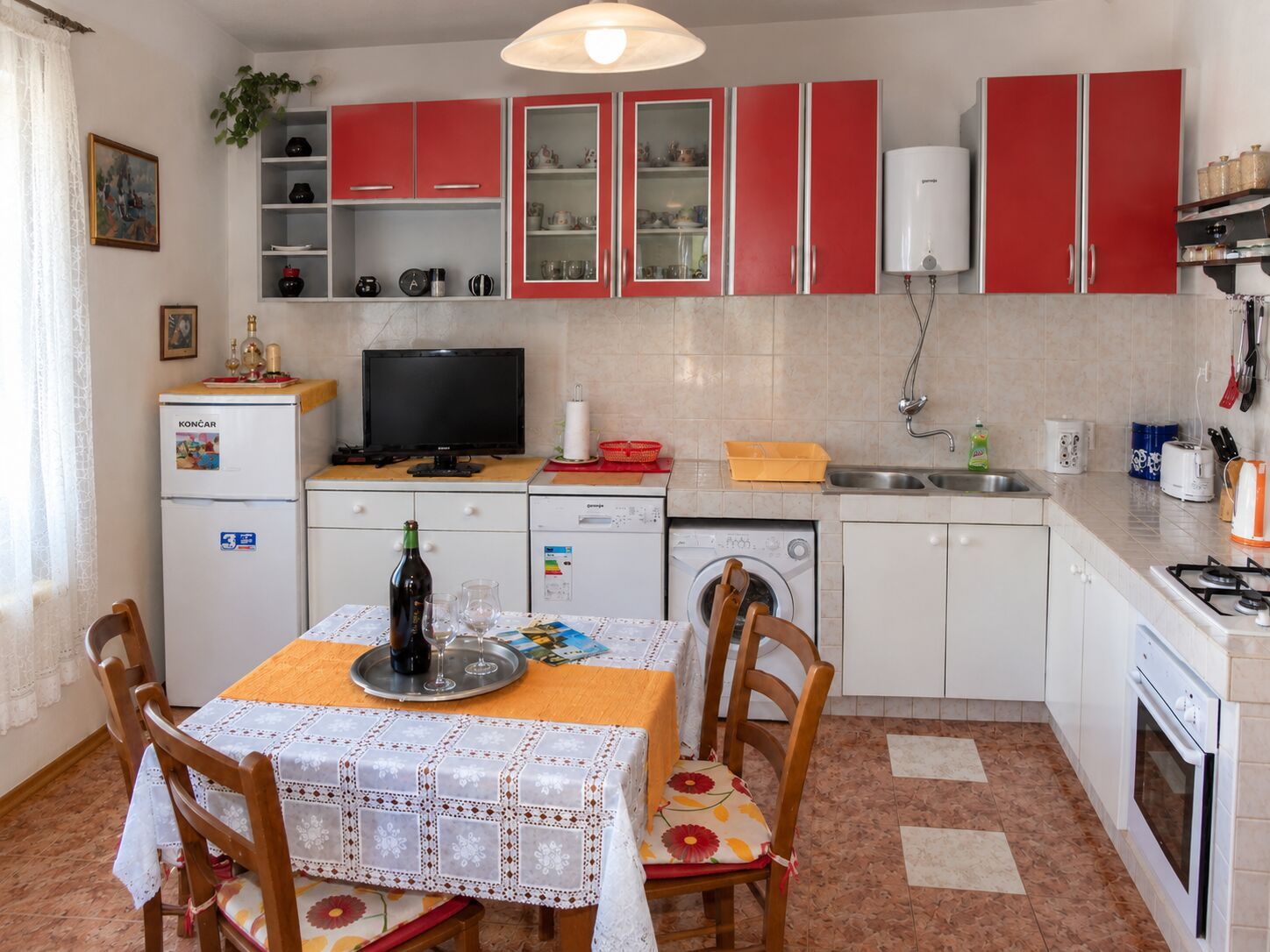 A compact kitchen with red cabinets, a dining table with chairs, a washing machine, and a small refrigerator.