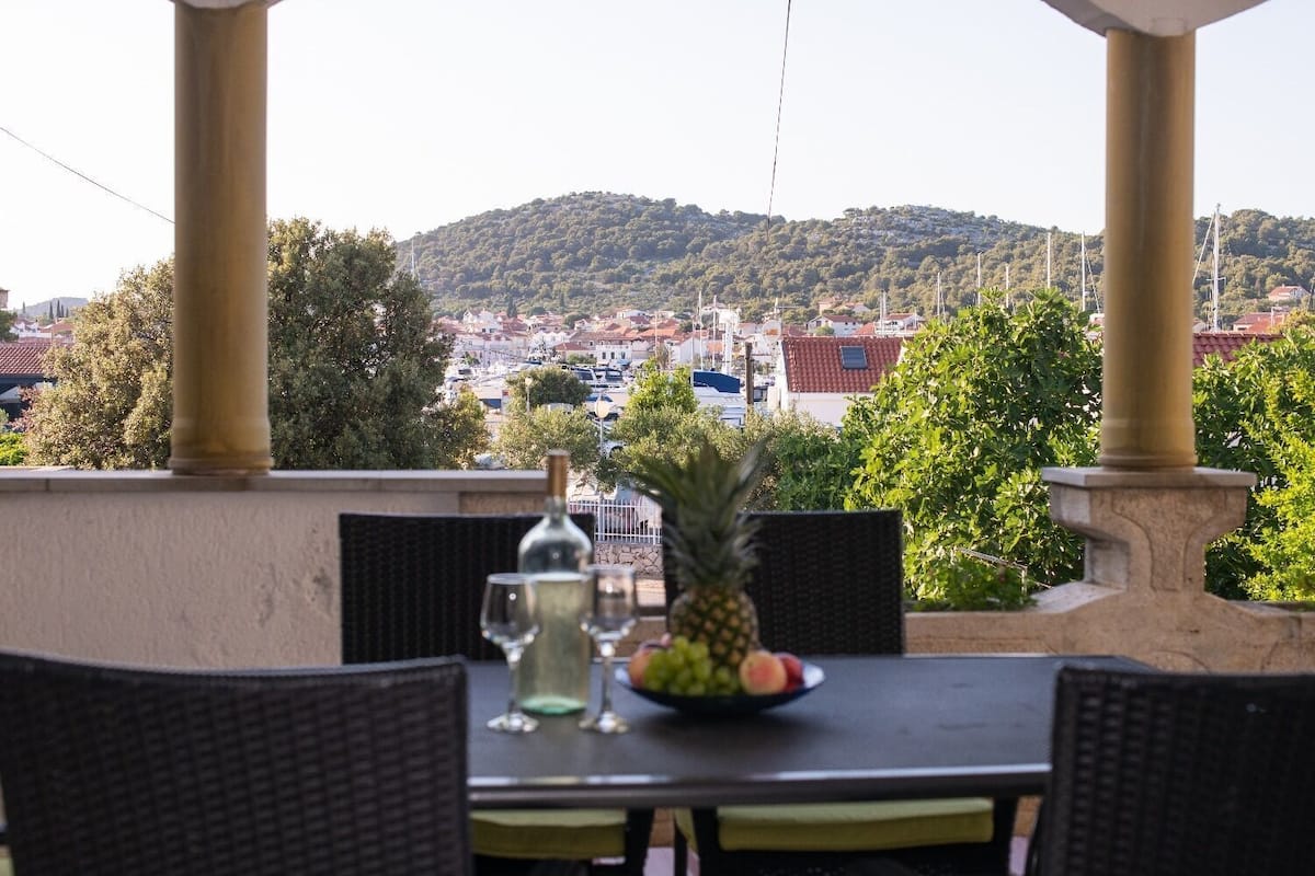 An outdoor dining area with a table set for two, a bottle of wine, and a pineapple. The setting includes wicker chairs and a view of a town with buildings and hills in the background.