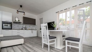 A modern kitchen with white cabinets, a built-in oven, and a marble backsplash. There is a dining area with a white table and chairs, and a large window with curtains. - Apartments Gabi i Miki (Trogir)