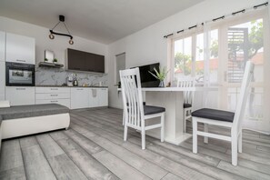 A modern kitchen with white cabinets, a built-in oven, and a marble backsplash. There is a dining area with a white table and chairs, and a large window with curtains. - Apartments Gabi i Miki (Trogir)