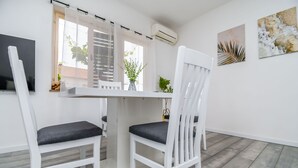 A dining area with a white table and chairs, a flat-screen TV mounted on the wall, and a window with curtains. - Apartments Gabi i Miki (Trogir)