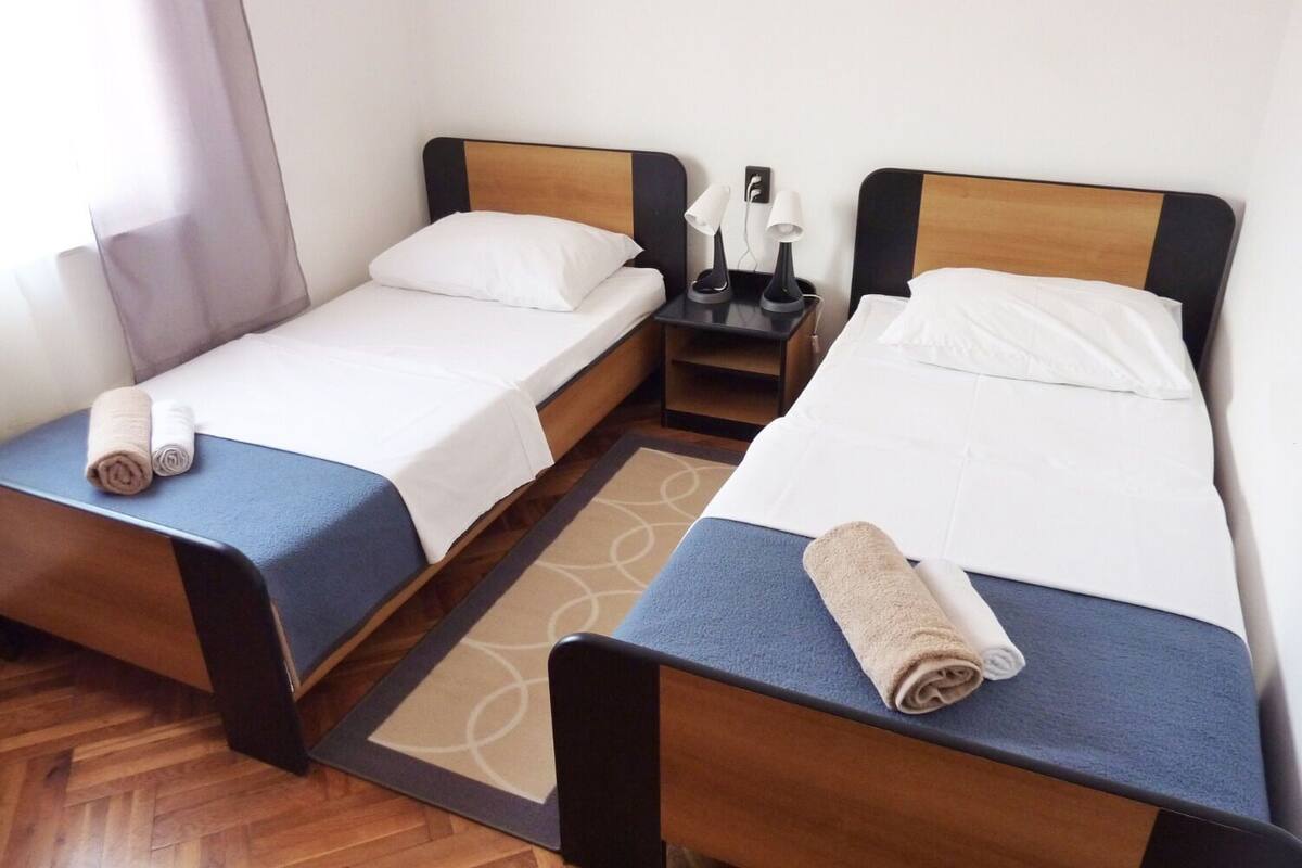 A hotel room with two single beds, each with a blue bedspread and white pillows, a small table with a lamp, and a wooden floor.