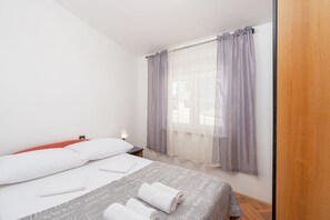 A neatly made bed with white pillows and a grey bedspread, a bedside table with a lamp, and a window with grey curtains. - Apartments Bozica (Vir)