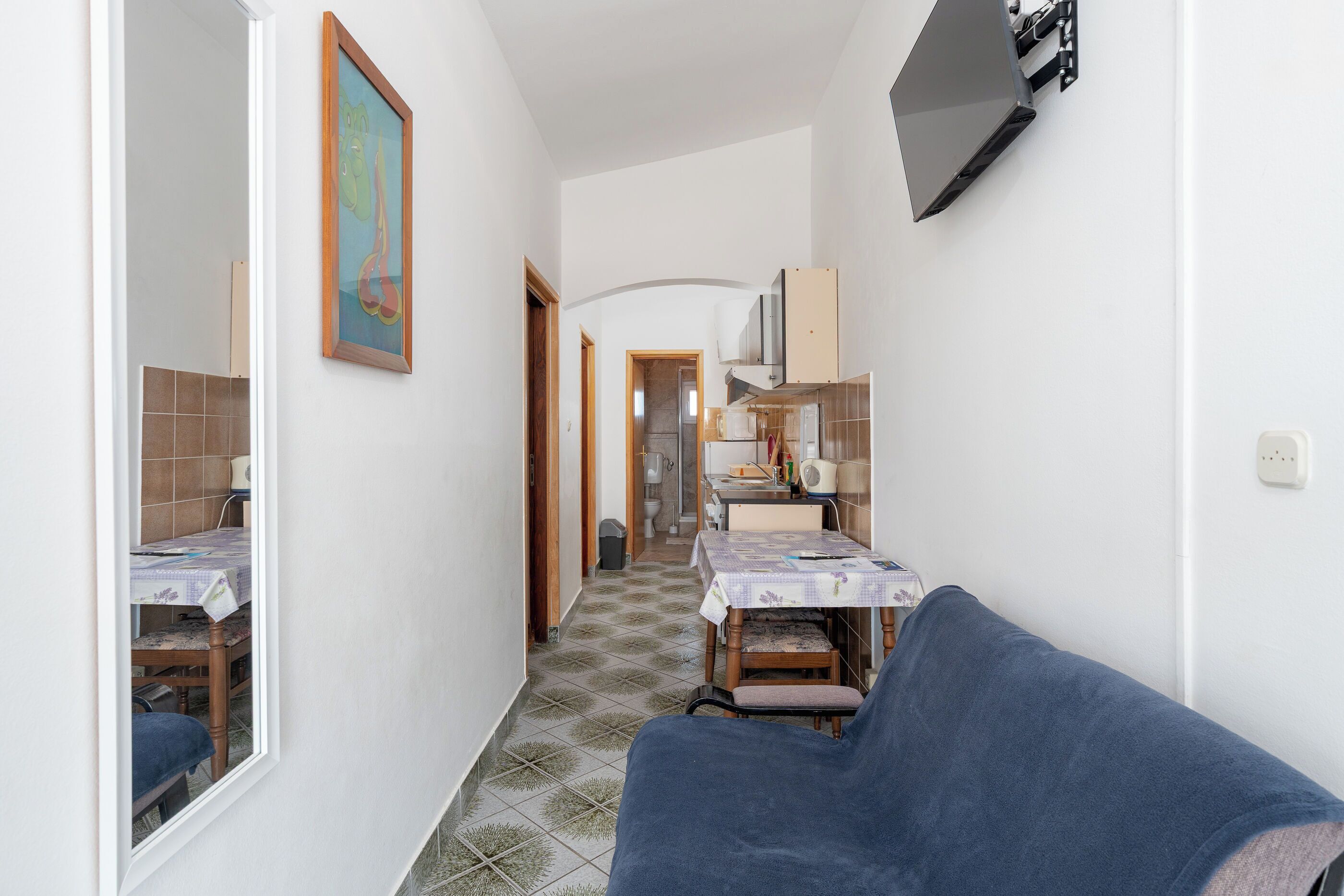 A narrow hallway with a dining area at the end, featuring a table and chairs, a blue sofa, and a painting on the wall.