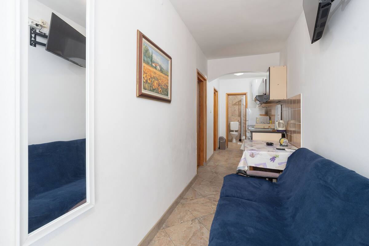 A narrow hallway with a blue sofa, a painting on the wall, and a kitchen area visible through an open door.