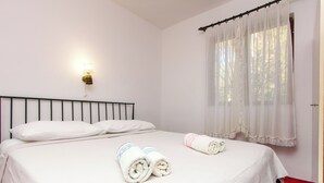 Una cama bien hecha con sábanas blancas y dos toallas enrolladas, una lámpara de pared y una ventana con cortinas translúcidas.