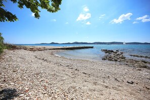 A pebbled beach with a pier extending into calm waters, surrounded by clear skies and distant hills. - Apartments Maja (Biograd na Moru)