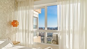 A hotel room with a bed, a window with a city view, a small table, and a wall-mounted lamp. - Apartment 'Top Costa Del Sol' with Shared Pool, Wi-Fi and Air Conditioning (Torremolinos)
