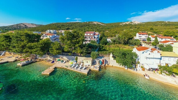 A coastal town with clear turquoise waters, a pier with sun loungers, and buildings with red-tiled roofs. - Apartments Jadra (Novalja)
