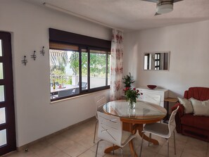 A dining area with a glass table, white chairs, and a vase of flowers. There is a window with a view of greenery and a red sofa in the corner. - House Igor (Rogoznica)