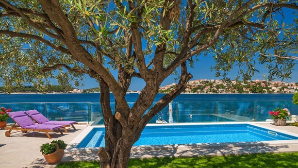 A pool area with a tree, lounge chairs, and a view of a coastal town. - House Lucmar (Rogoznica)