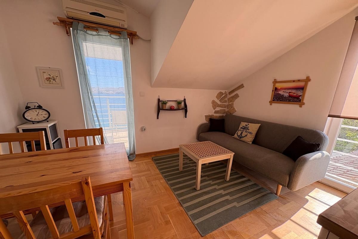 A cozy living room with a wooden dining table, a sofa, and a small coffee table. There is a clock on the wall and a window with a view.