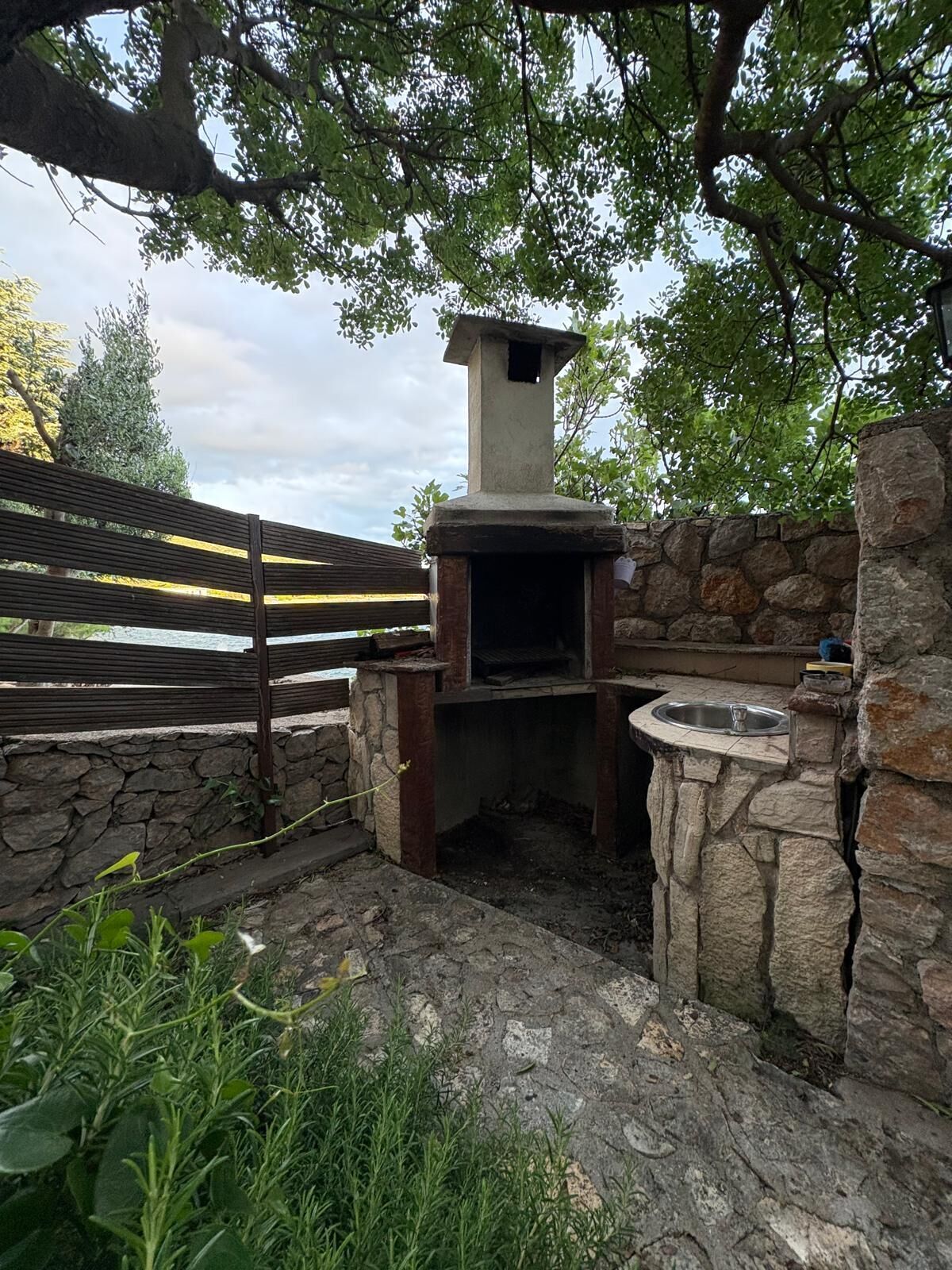 An outdoor stone and wood-built barbecue area with a stone chimney, surrounded by greenery and a stone wall.