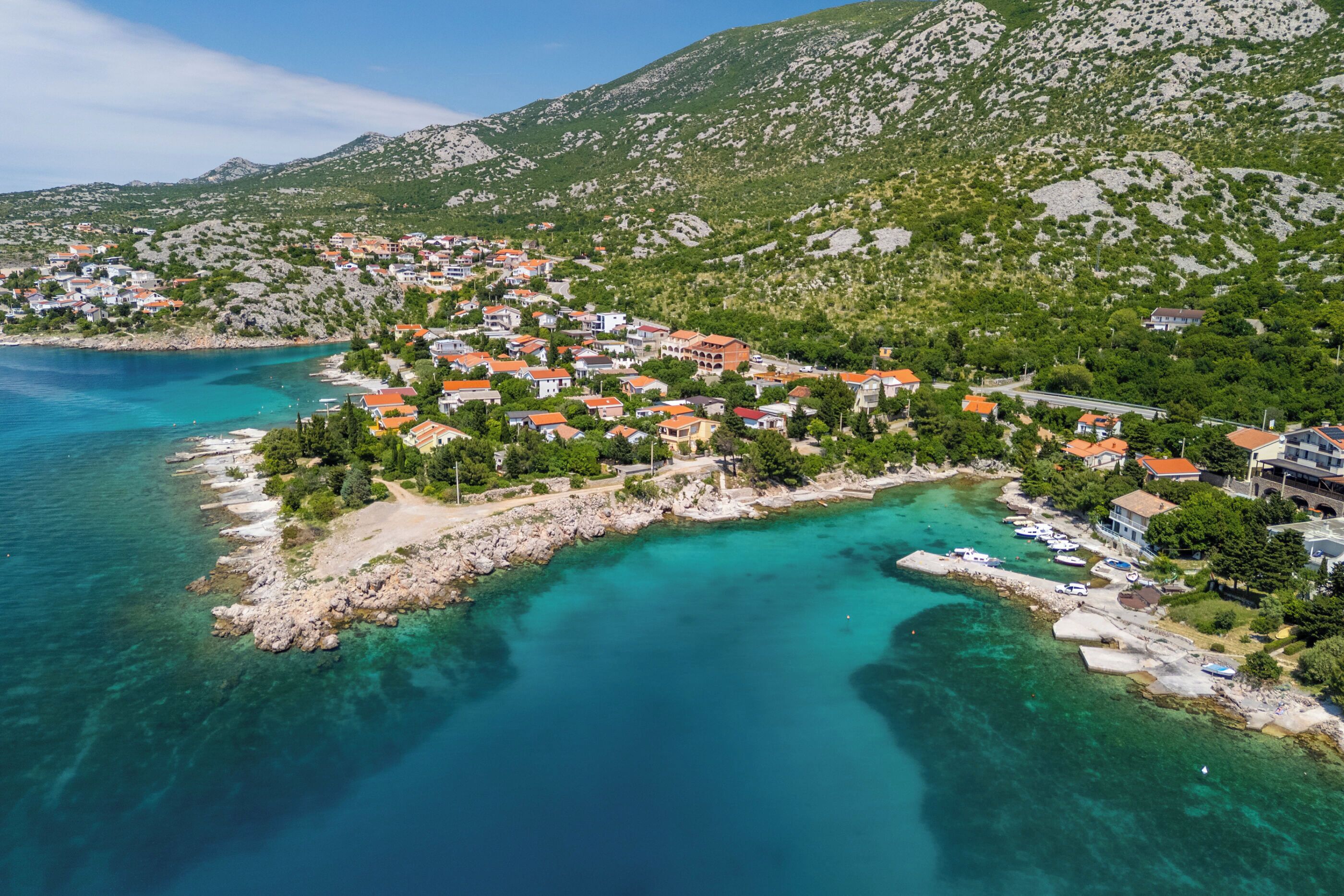 Eine Küstenstadt mit Häusern entlang der Uferlinie, klarem türkisblauem Wasser und grünen Hügeln im Hintergrund.