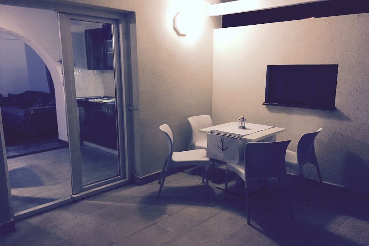 A small, modern room with a glass door leading to a balcony, a white table with chairs, and a mounted TV on the wall.