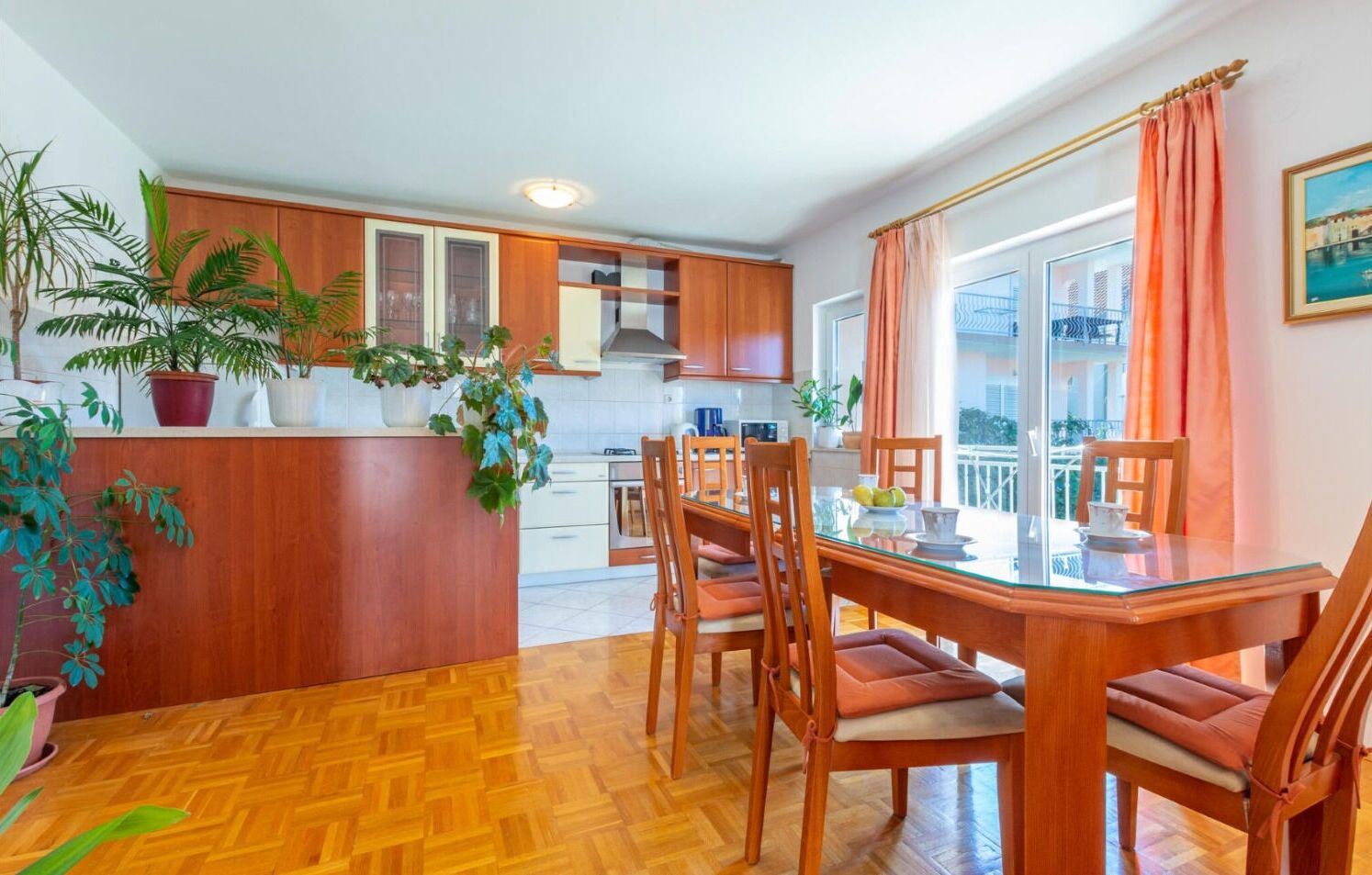 A modern kitchen with wooden cabinets, a dining area with a glass table and chairs, and potted plants.