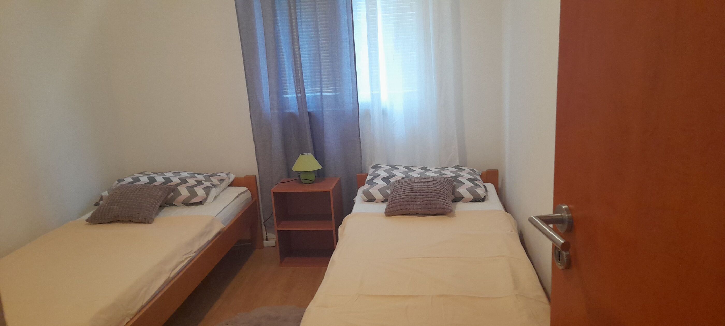 Une petite chambre propre, avec deux lits simples, une table de chevet avec une lampe et une fenêtre ornée de rideaux légers.