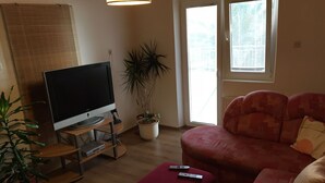 Un salon avec un téléviseur à écran plat posé sur un meuble, un canapé rouge, une plante en pot et une fenêtre avec des stores.