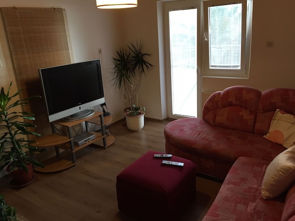 A living room with a flat-screen TV on a stand, a red sofa, a potted plant, and a window with blinds. - Apartments Durda (Pirovac)