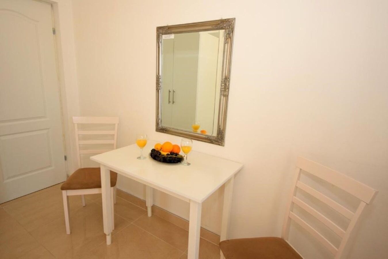 Um quarto pequeno e branco, com uma mesa branca e duas cadeiras. Um espelho está pendurado na parede acima da mesa, e há uma fruteira com laranjas sobre a mesa.