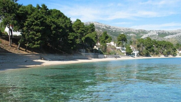 Ein Strand mit klarem Wasser, sandigem Ufer und Häusern im Hintergrund.