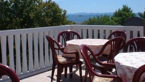 Eine Terrasse im Freien mit runden Tischen und Stühlen, einem weißen Zaun und Blick auf das Meer und die Berge im Hintergrund.