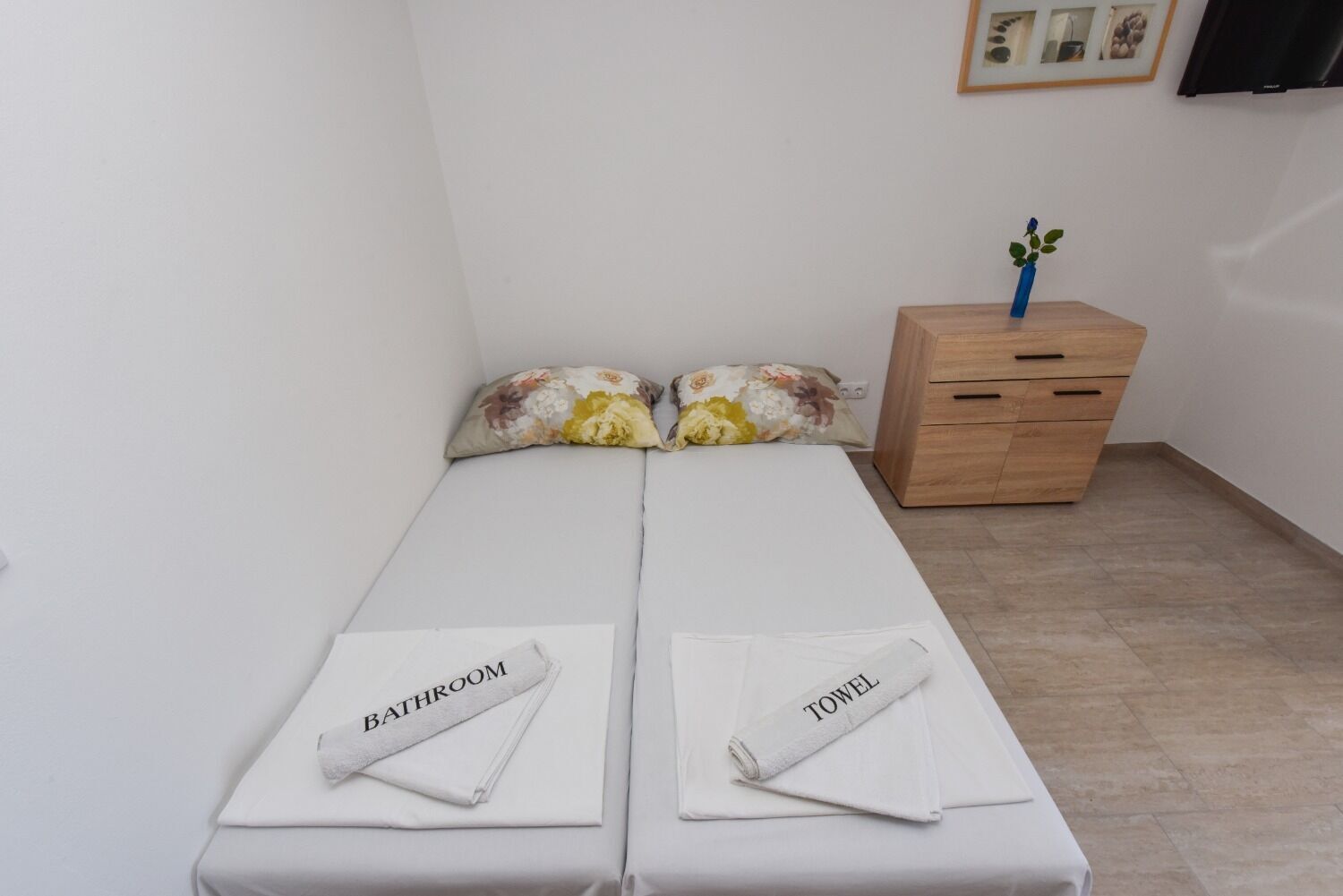 Une petite chambre propre, avec un lit, deux oreillers, une table de chevet en bois ornée d’un vase et un tableau encadré accroché au mur.
