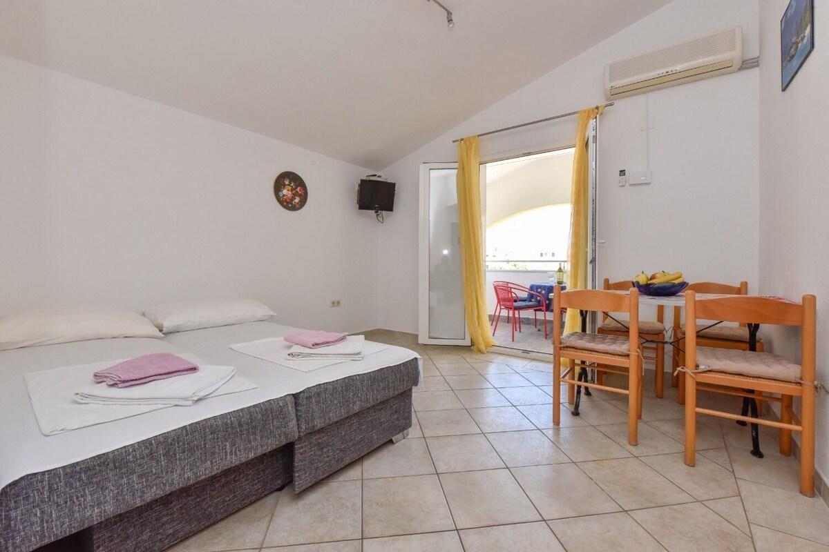 A bedroom with a bed, a desk with chairs, a window with curtains, and an air conditioning unit.