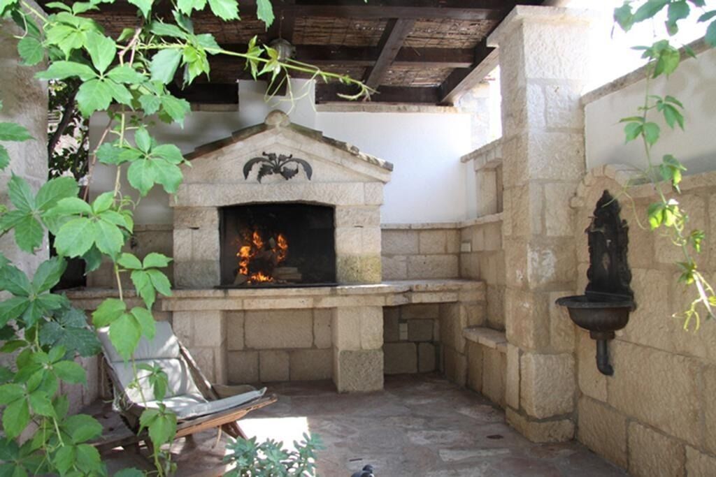 A stone-built outdoor fireplace with a fire burning, surrounded by stone walls and a rustic wooden ceiling.