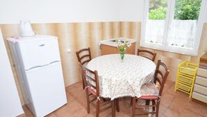 Eine kleine Küche mit einem weißen Kühlschrank, einem runden Tisch mit einer blumigen Tischdecke und Holzstühlen. Ein Fenster mit Spitzenvorhängen bietet Blick auf grüne Vegetation.