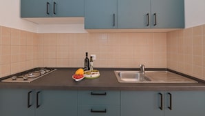 A modern kitchen with teal cabinets, a gas stove, a sink, and a countertop with a bowl of fruit and a bottle of olive oil. - Apartments Ilija (Novalja)