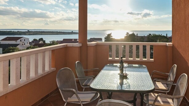 A balcony with a glass table and chairs, overlooking a body of water and distant land. - Apartments Ilija (Novalja)