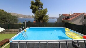 Una piscina con vistas al mar y a las colinas, rodeada por una valla y una casa con techo rojo.