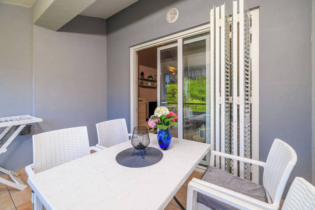 A white table with four chairs, a vase with flowers, and a sliding glass door leading to a balcony.
