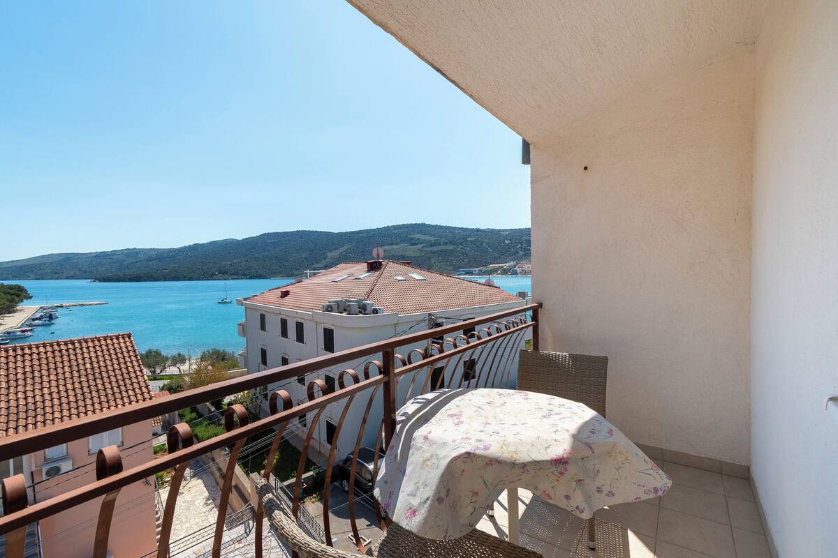 A balcony with a view of a coastal town, a table with a floral tablecloth, and a metal railing.