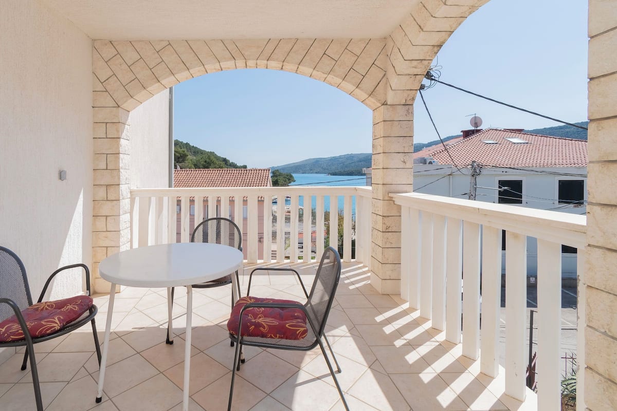 A balcony with a round table and two chairs, a view of the sea and hills, and a white railing.