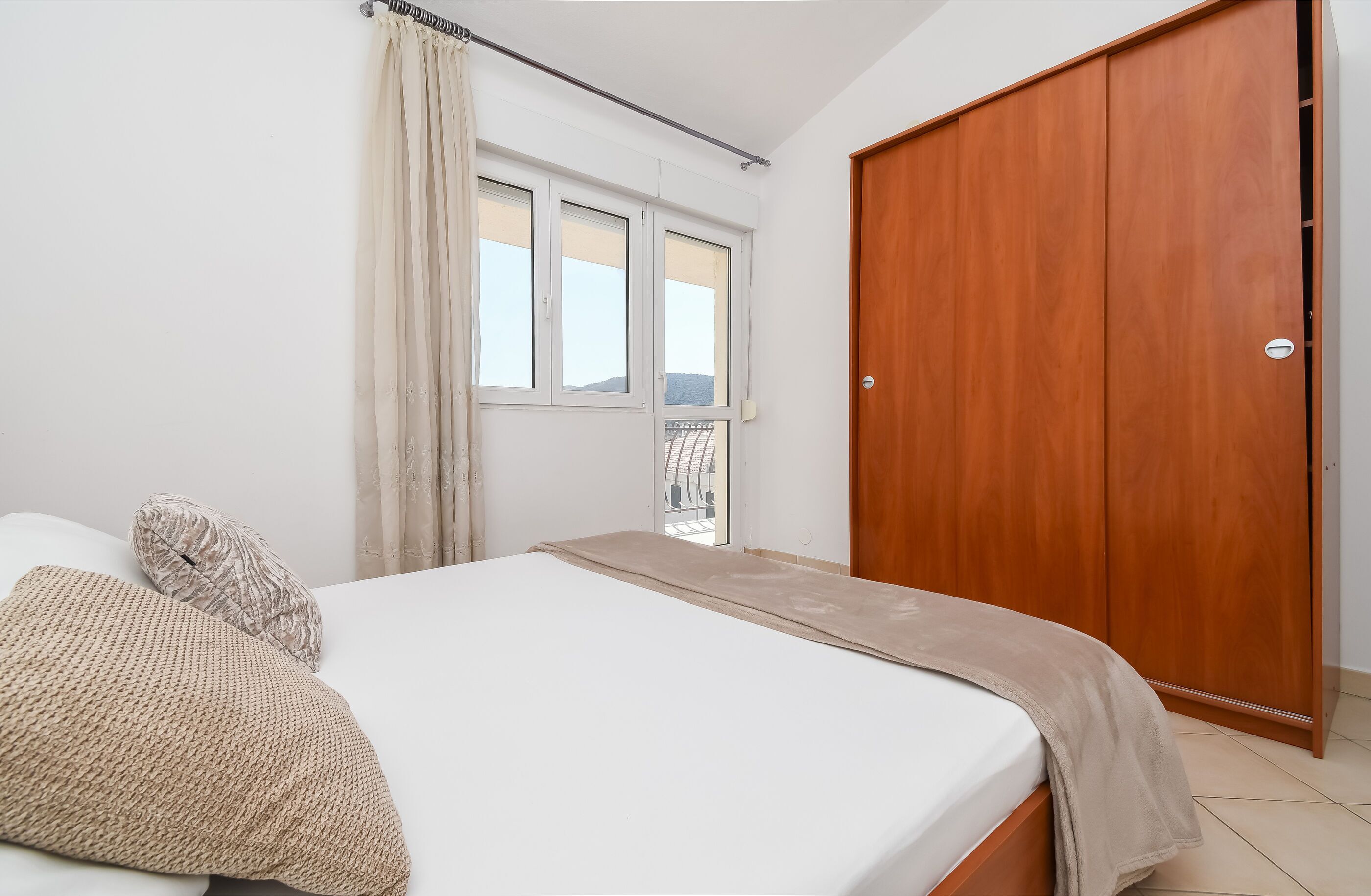 A bedroom with a bed, a wooden wardrobe, a window with curtains, and a view of a balcony.