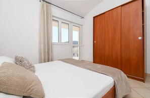 A bedroom with a bed, a wooden wardrobe, a window with curtains, and a view of a balcony. - Apartments Pera (Marina)