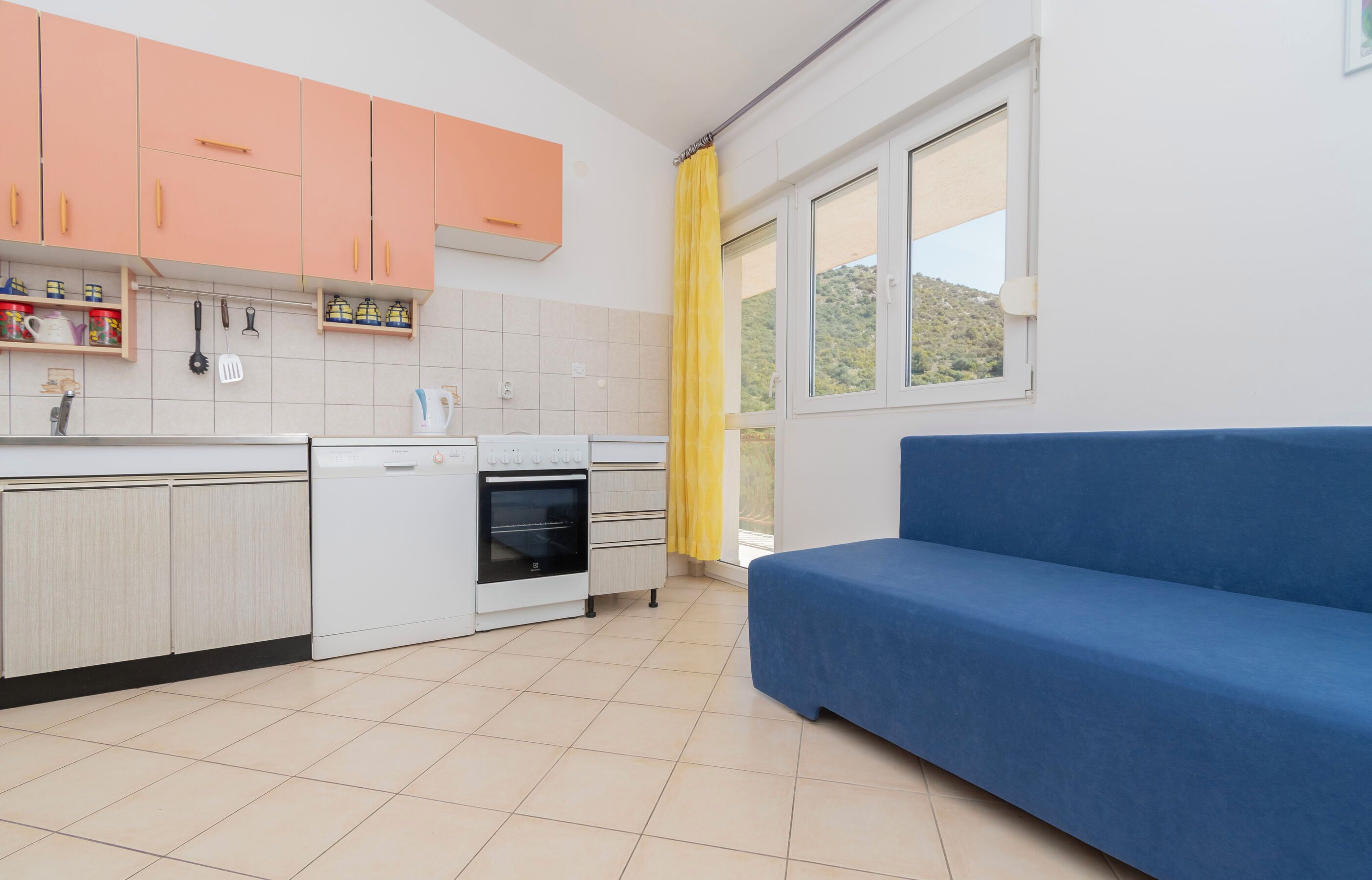 A modern kitchen with white cabinets, a blue sofa, and a window with a yellow curtain.