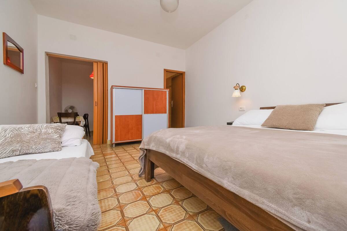 A hotel room with two beds, a wooden wardrobe, a small table with chairs, and a wall-mounted lamp.