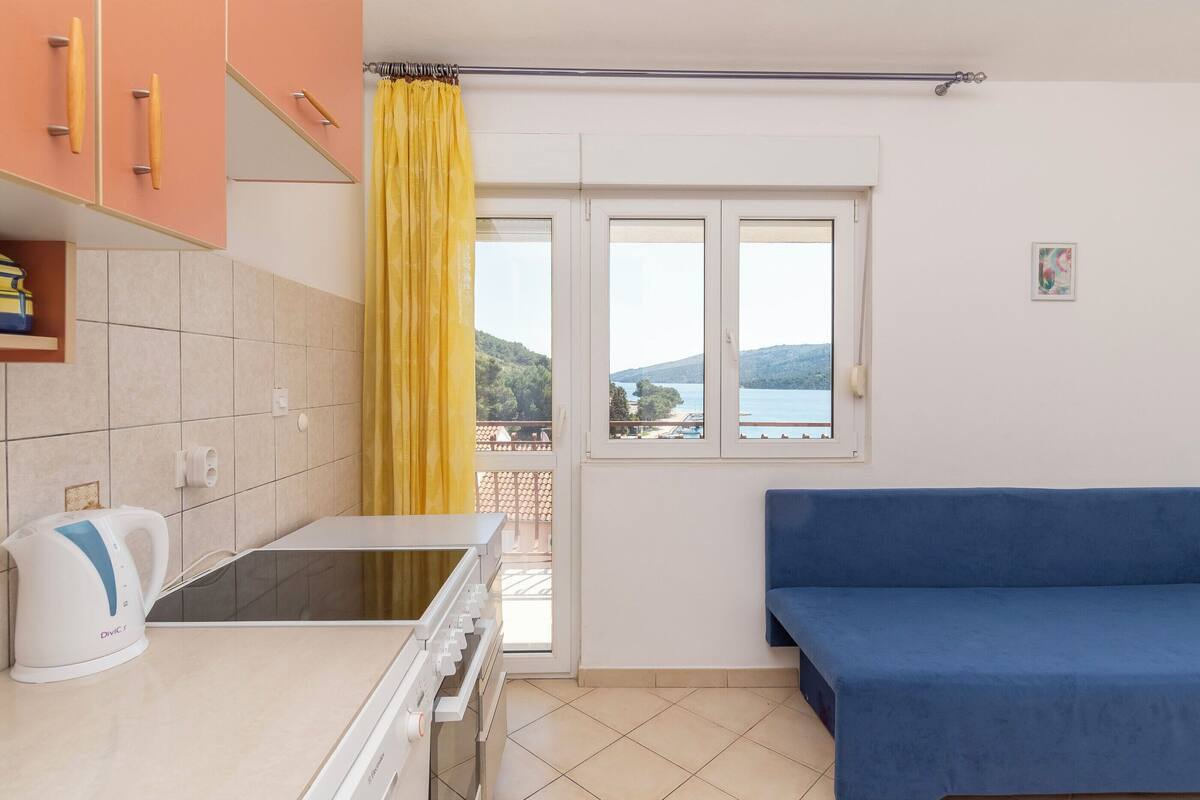 A small, bright room with a kitchenette, a blue sofa, and a window with a view of a lake and hills.
