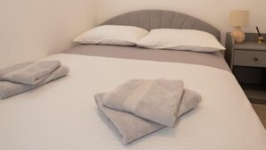 A neatly made bed with folded towels, a bedside table with a lamp, and a window with blinds. - Apartment Danka (Novalja)