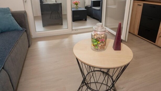 A modern living room with a grey sofa, a round coffee table with a glass jar and a purple vase, and a sliding glass door leading to a balcony with outdoor furniture. - Apartment Danka (Novalja)