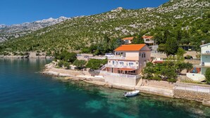 A coastal building with a red roof, situated by a clear blue sea, with mountains in the background. - Apartment Dalibor (Karlobag)