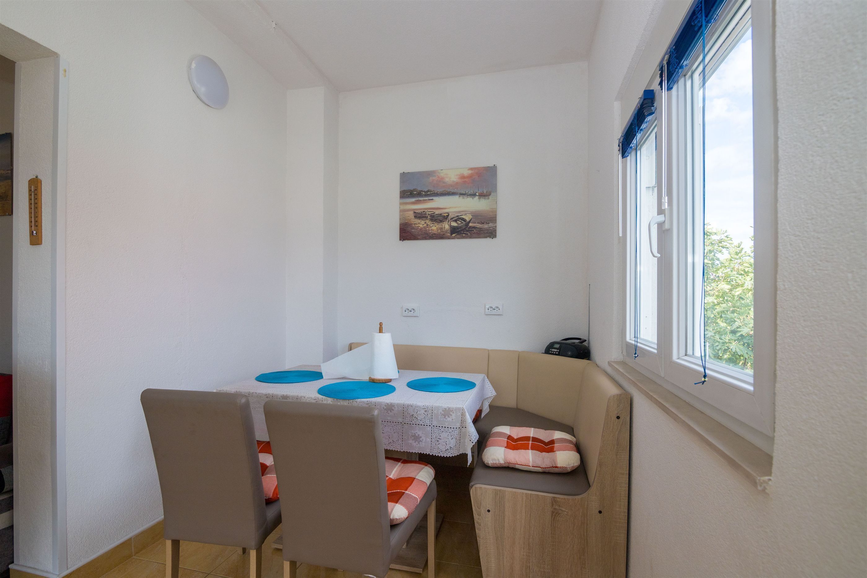 Un petit coin repas avec une table et des chaises, un tableau au mur et une fenêtre avec des stores.