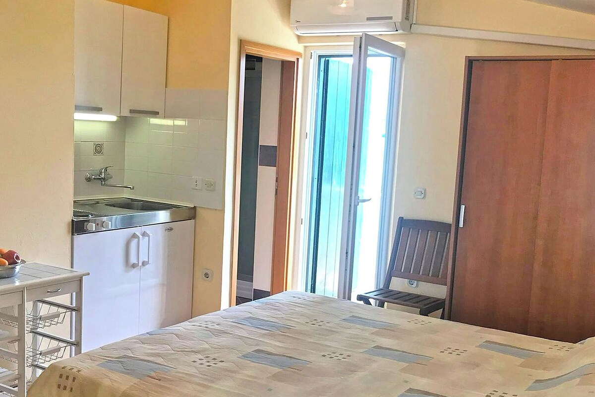 A compact bedroom with a bed, a small kitchenette, and a sliding door leading to a balcony.