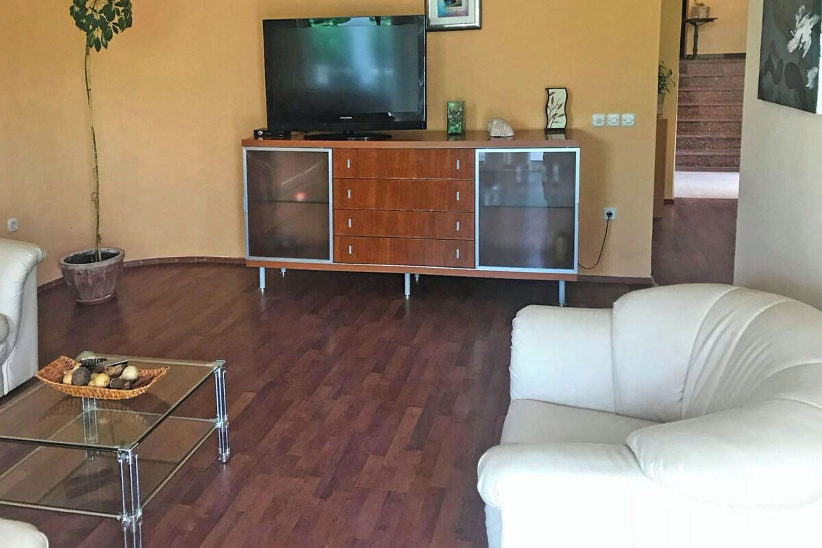 A living room with a flat-screen TV on a wooden cabinet, a potted plant, a glass coffee table, and white sofas.