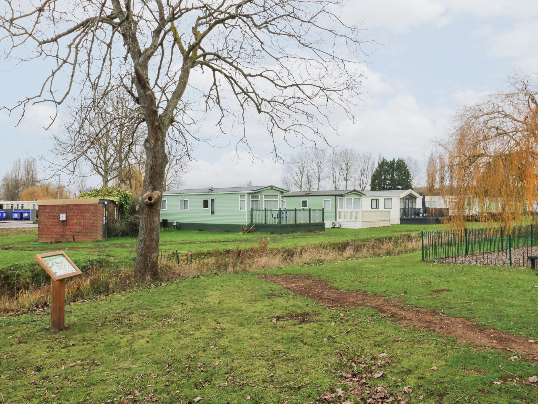 A row of mobile homes with a grassy area in front, a tree, and a small brick structure.