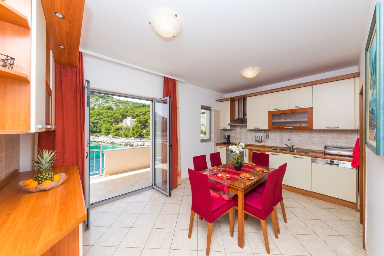 A modern kitchen with a dining area, featuring a large table with red chairs, a view of the sea, and a balcony with a sliding door.