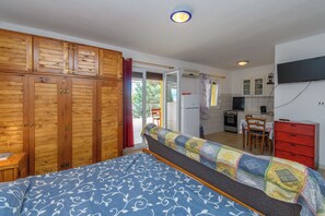 A bedroom with a bed, wooden wardrobe, and a kitchenette in the background.